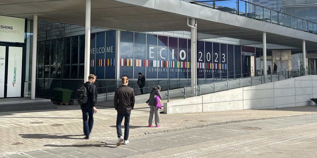 Physical advertising spaces Visitors walk towards the entrance of a conference center with large signs welcoming them to ECIO 2023, an international congress on oncological interventions.