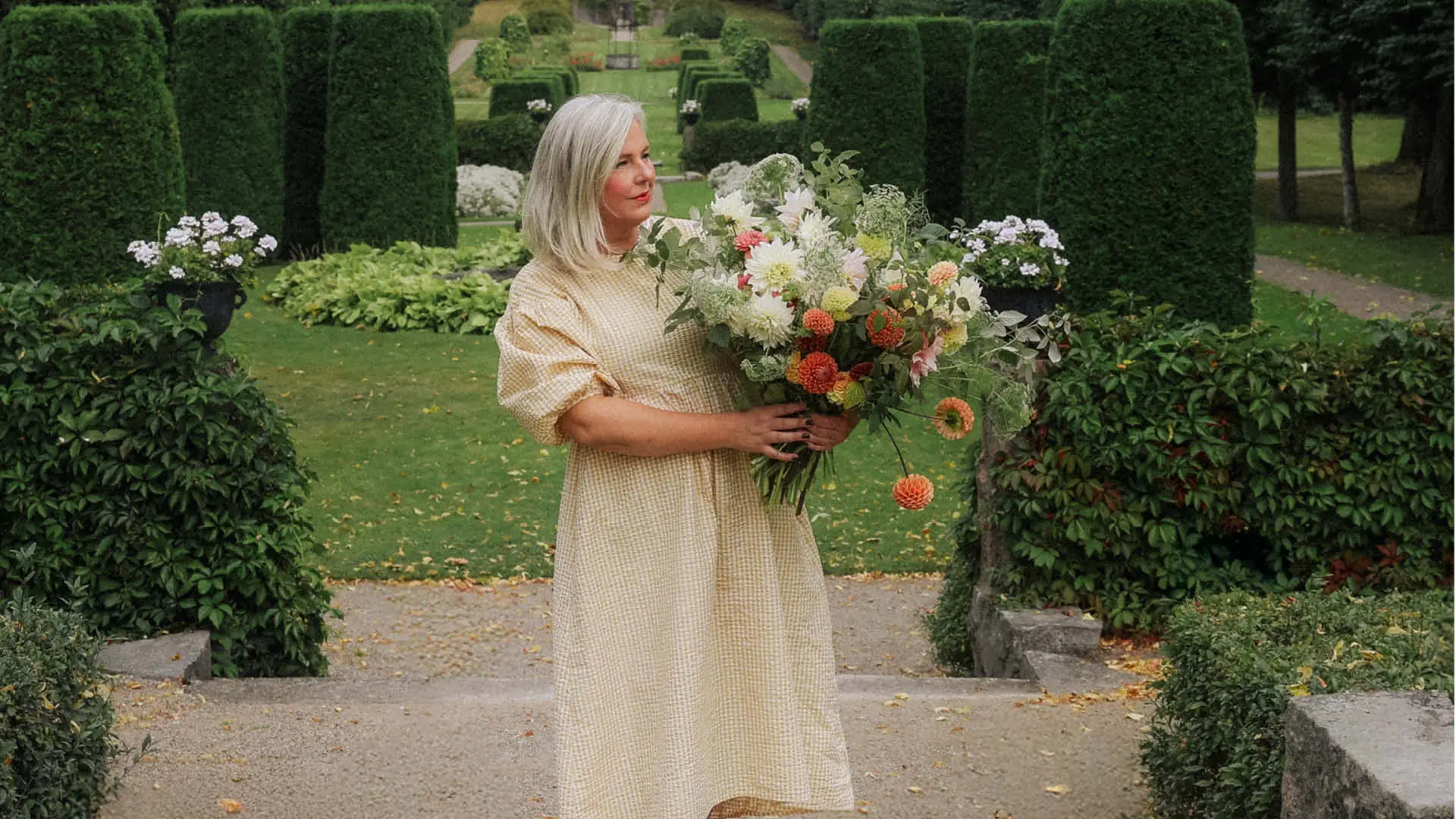 webb-workshoplshanna (1) En kvinna i klänning står i en park med ett fång blommor i famnen